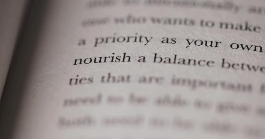 The camera focuses on an open book displaying two lines of text a priority as your own nourish a balance betw the surrounding words on the page are blurred
