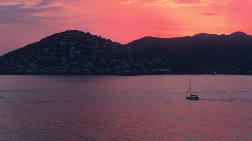 A sailboat glides peacefully across calm water as the sun sets casting a vibrant pink and orange glow behind a large dark mountain