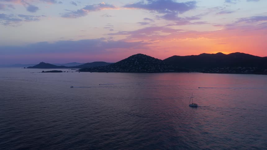 A beautiful aerial shot captures sailboats on the calm ocean as the sun sets behind silhouetted mountains painting the sky in vibrant colors