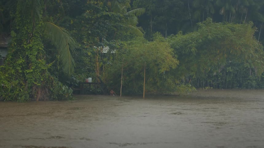 A wide shot of a flooded tropical area with muddy water covering the ground and a person walking through the water near the edge of a cluster of trees and buildings