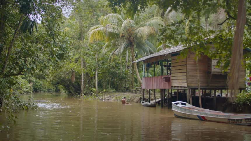A person swimming in a river near the bank with a shack and a canoe beside it surrounded by trees