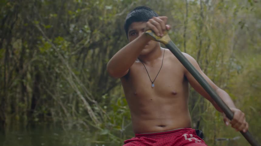 A young man with dark hair shirtless and wearing red shorts propels himself forward with a paddle in a calm body of water surrounded by lush foliage