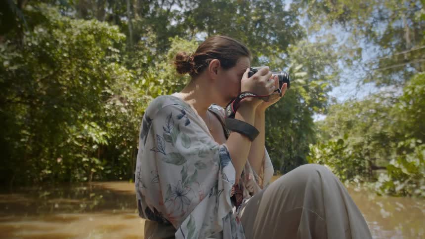 A person sits on a boat in a jungle setting taking photos with a camera they are surrounded by lush green foliage and are wearing a lightcolored patterned kimonostyle top