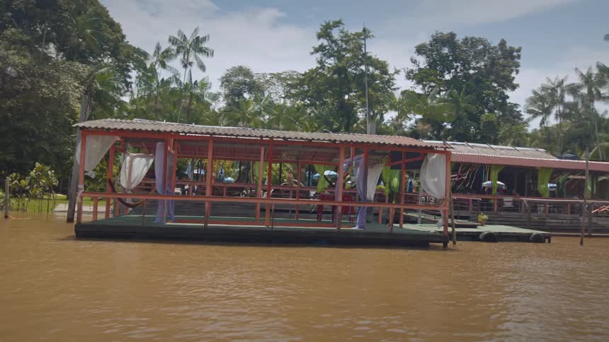 Two floating restaurant structures with corrugated metal roofs and light wooden frames with white and green curtains hanging from the eaves moored to a muddy brown riverbank covered in dense green jungle and palm trees under a cloudy sky