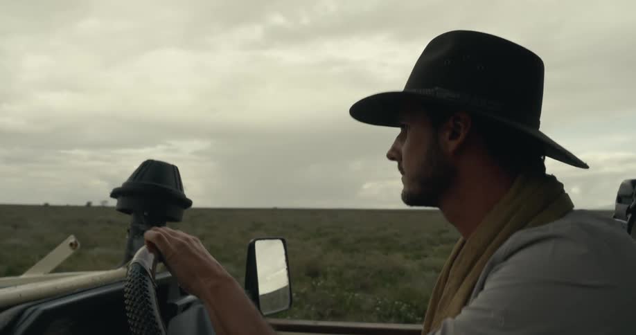 The video shows a man wearing a widebrimmed hat and a scarf driving a vehicle through a grassy landscape he is focused on the road ahead and the sky is overcast