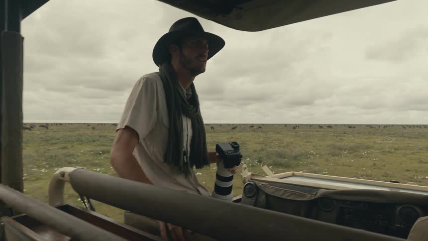 Man in a safari vehicle holding a camera with a long lens while watching animals on the savannah