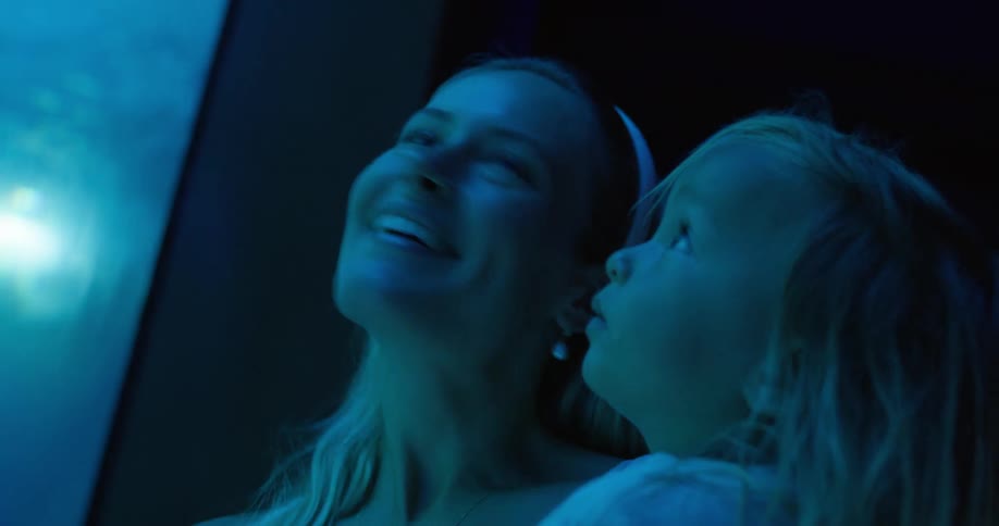 A woman with long blonde hair and a young child with long blonde hair stand together in a dimly lit aquarium with them smiling and pointing up
