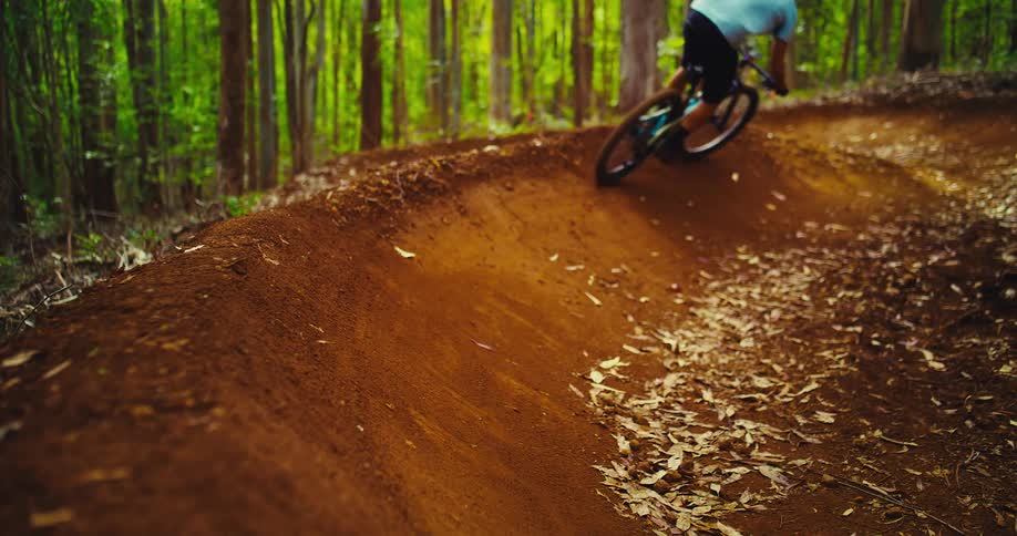 The video shows a mountain biker riding on a dirt track through a forest the camera follows close behind as the bike goes around a bend