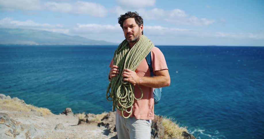 A man holding a bundle of climbing ropes over his shoulder carrying a backpack and standing on a cliff by the shoreline at midday