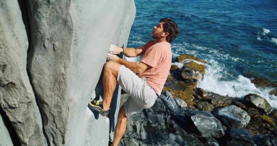A man with white chalk in his hands is climbing a large rock on a coast with sea waters breaking below