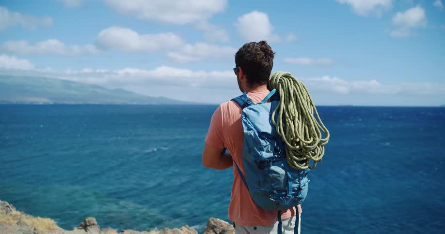 A man holding a bundle of climbing ropes over his shoulder carrying a backpack and standing on a cliff by the shoreline at midday