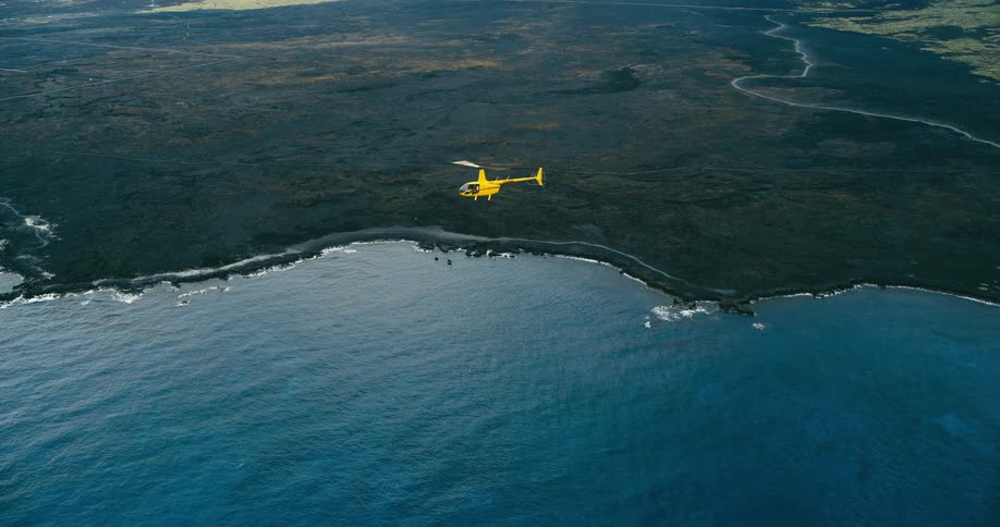 A bright yellow helicopter flies above a rugged shoreline and blue water