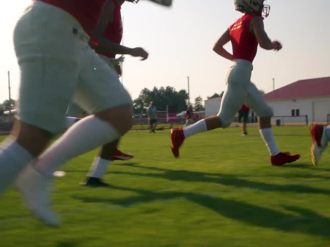 A football team in red jerseys and white pants is practicing on a grass field under a clear sky they run in a line from a crouched position toward the left side of the frame