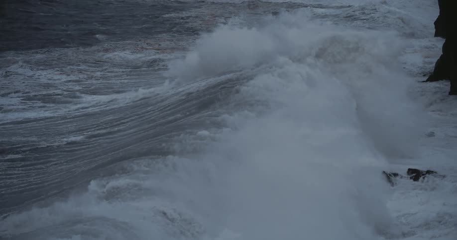 A very large wave with white foam breaks against a dark rocky cliff causing a large splash of white water to shoot up the side of the cliff