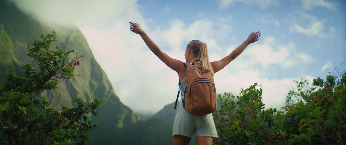 A young woman standing on a cliff with both hands raised in excitement during a hike carrying a backpack at midday with a mountain view in the distance