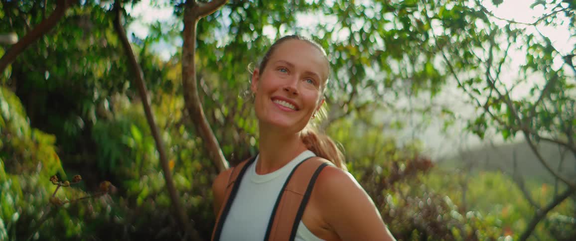 Smiling woman with backpack poses on a trail lit by the sun among trees