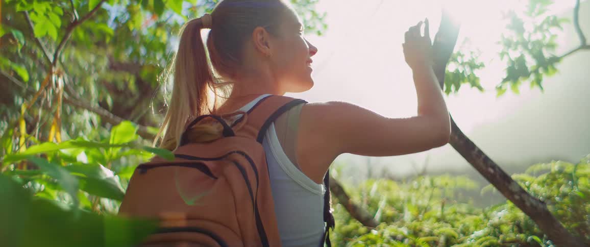 A blonde woman with a backpack is walking through a bright green lush forest she grabs onto a tree branch and then continues on her path