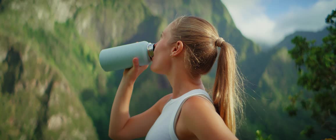 A woman in athletic wear drinks water with mountains behind