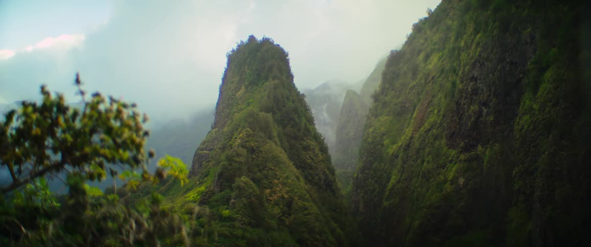 Mist drifts through a valley between majestic junglecovered mountains revealing a thin waterfall in the distance