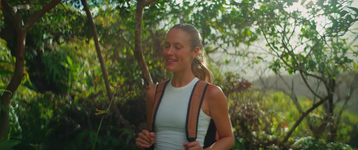 A woman with a backpack and a white tank top stands among trees looking up and taking a deep breath before turning to face the camera and smiling all while the bright sun shines through the leaves creating a lens flare