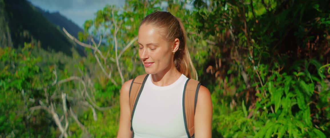 A woman with her hair in a ponytail and a white tank top looks from side to side and smiles as she walks in a bright forest with a tan backpack on her back