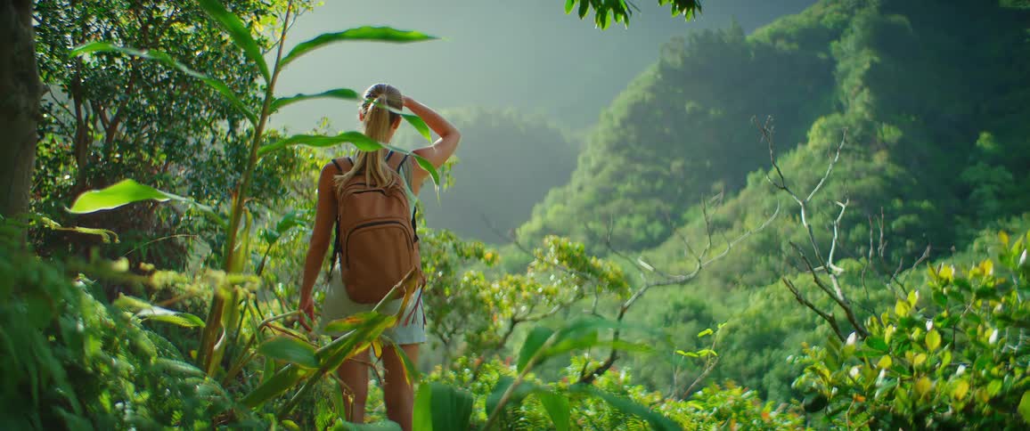 A woman with a backpack and a ponytail stands on a trail surrounded by lush green foliage looking out over a misty mountain landscape