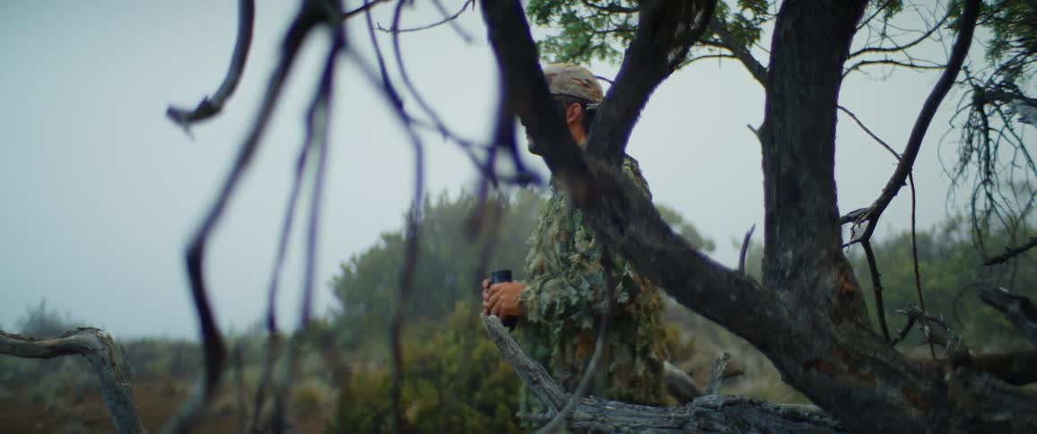 A man in a ghillie suit uses binoculars to look into the distance he is standing behind a tree with a branch in the foreground