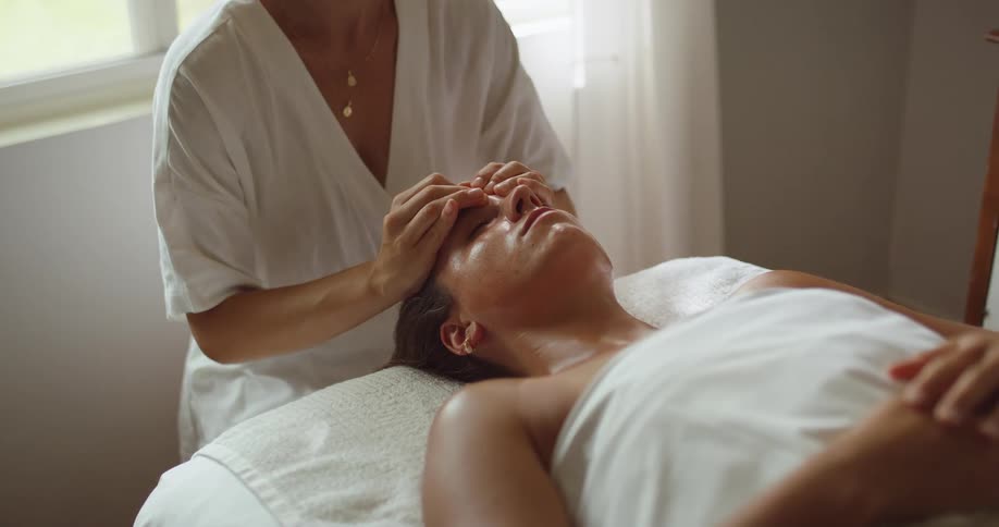 Woman lies on a massage table while a massage therapist massages her face during the daytime