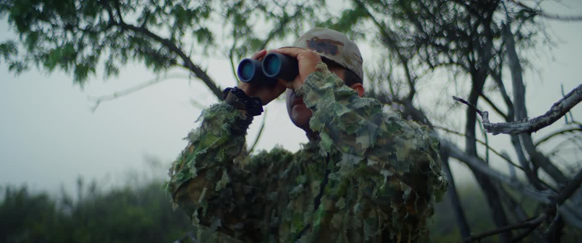 A person wearing a camouflage jacket and hat raises a pair of binoculars to their eyes they are standing in a wooded green area