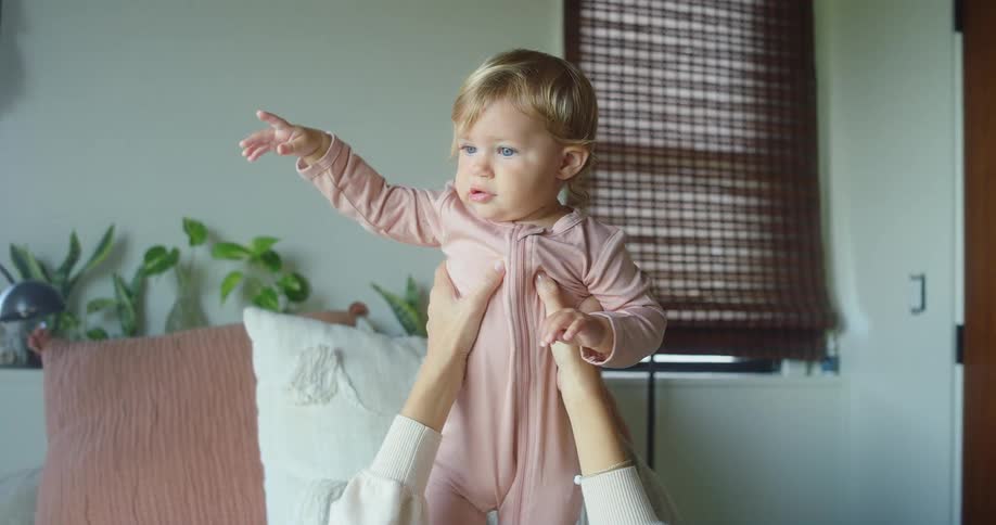 Baby in pink onesie is lifted by hands indoors