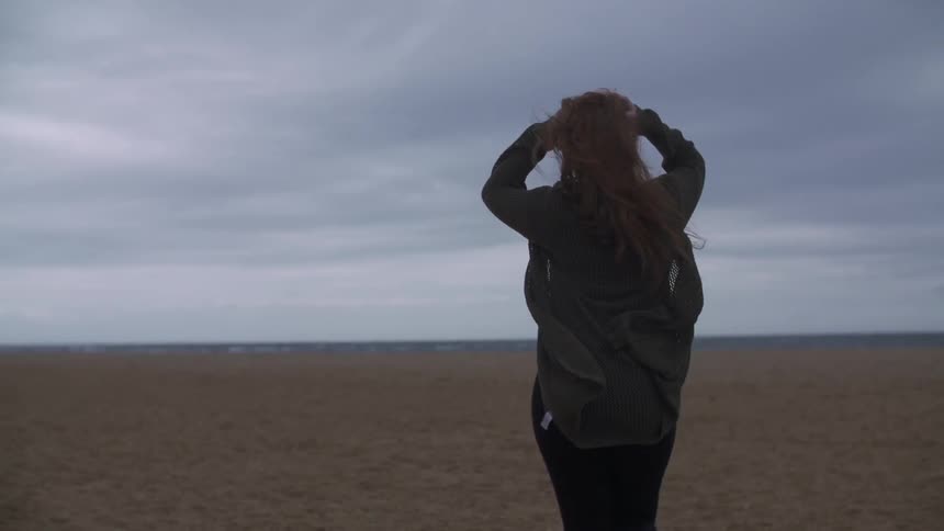 A woman is walking on the beach while the wind blows her long light brown hair