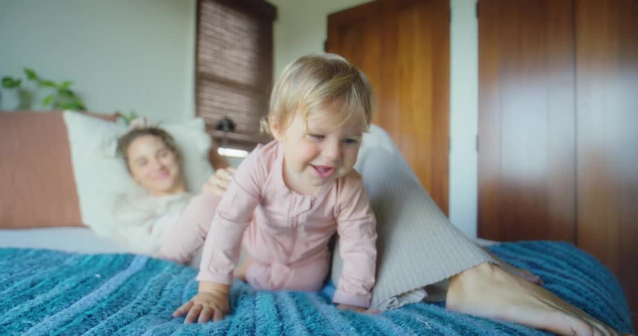 A toddler crawls on a bed as a smiling mother watches