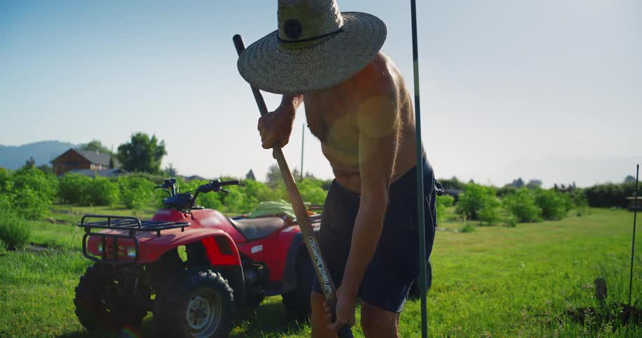 A man wearing a straw hat and black shorts with no shirt on walks across a grassy field using a shovel to fill a hole while a red atv and a trailer are visible behind him