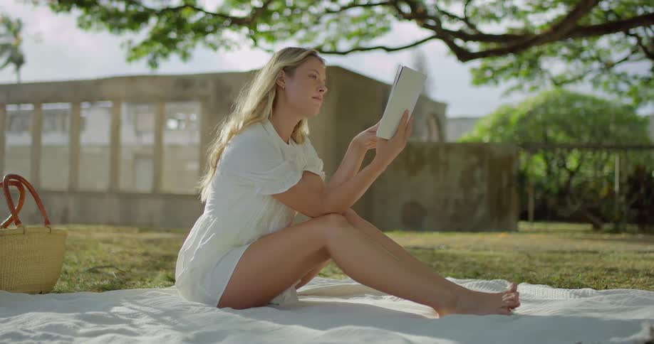 The video shows a wide shot of a young woman sitting on a white blanket in a park she is reading a book and a straw bag is visible beside her the background shows a building and trees suggesting a peaceful sunny day