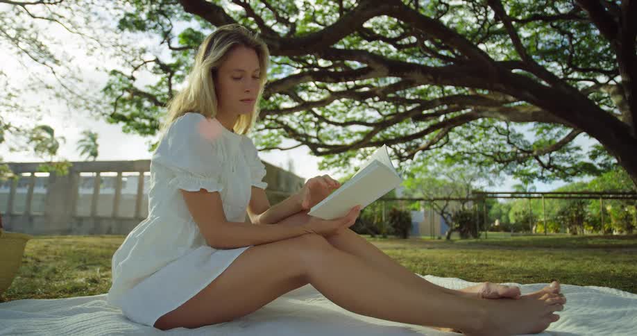 A young woman sits barefoot on a blanket under a large tree reading a book she pauses looking up and off into the distance