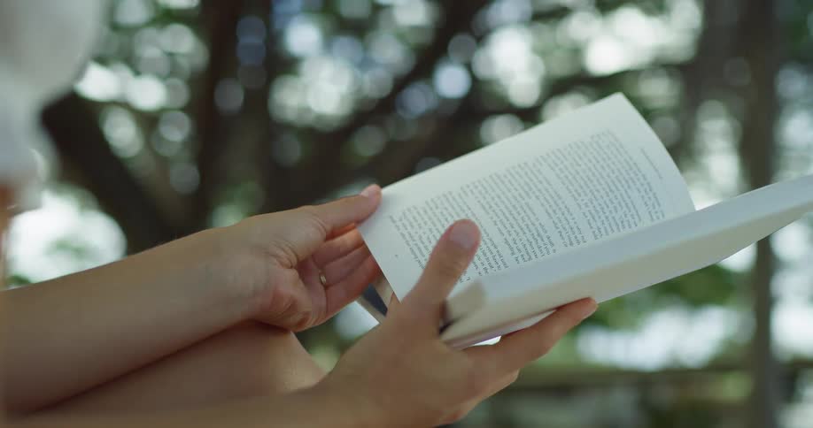 The video shows a closeup shot of a persons hands as they hold and read a book outdoors the pages flutter in a light breeze and the background is a blurry mix of green leaves and sunlight the watermark filmsupply is visible on the video