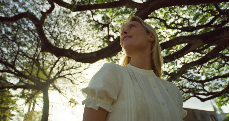 The woman walks under the tree gazing at the branches above