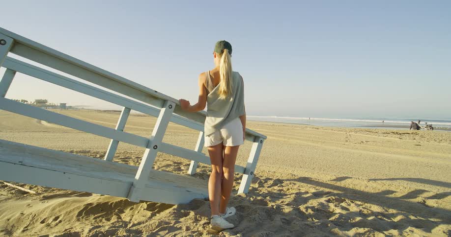 Woman in cap looks at the sea leaning on a wooden structure in the sand