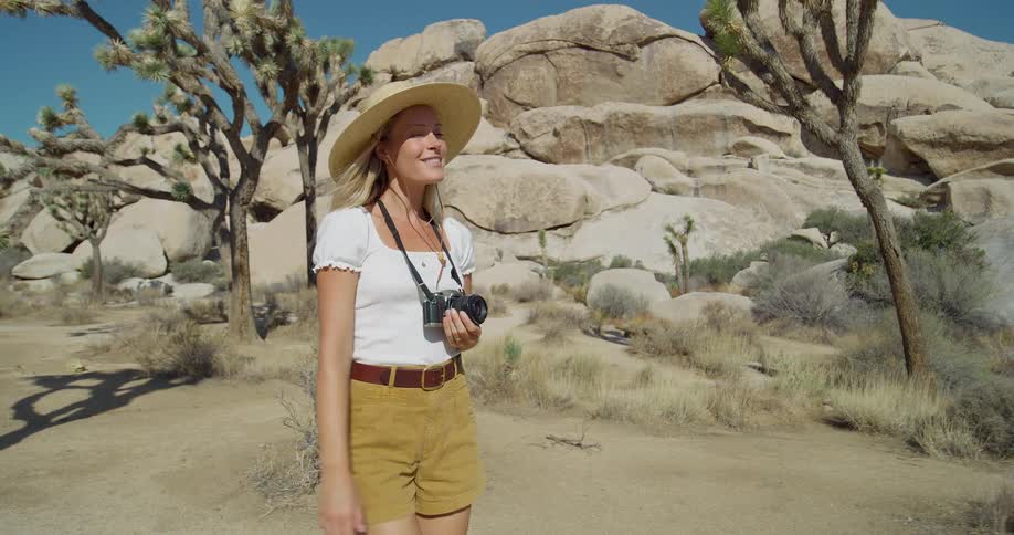 A woman wearing a straw hat and holding a camera walks through a desert landscape with joshua trees and large rock formations