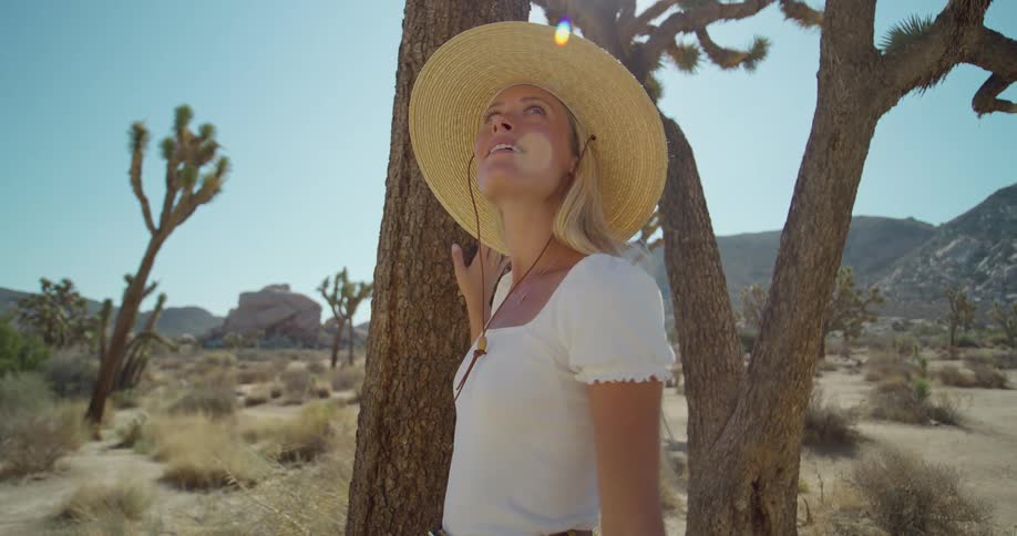 A woman with blonde hair wearing a straw hat and a white shirt stands next to a joshua tree looking up at the bright sun and smiling