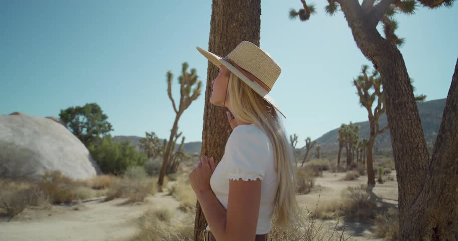 The video shows a woman in a widebrimmed straw hat standing in a desert landscape with joshua trees she is seen from the side as she turns her head to look at something in the distance