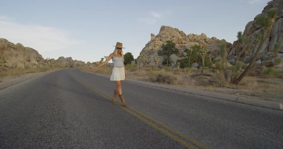 Woman in a white dress and hat walking in the middle of a deserted road surrounded by rocks and joshua trees