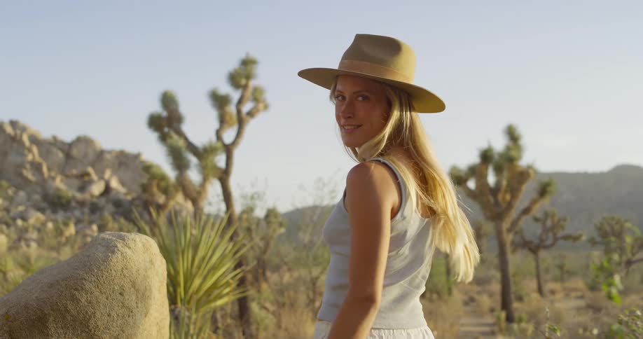 A young woman wearing a hat walks through a barren land filled with tough plants on a sunny day smiling