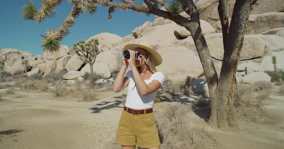 A blonde woman wearing a widebrimmed straw hat and a white shirt with brown shorts stands in a desert she holds a camera and smiles as she looks into the distance then brings the camera up to her eye to take a picture