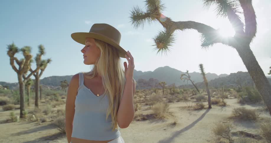 A woman with blonde hair wearing a wide brim hat and a light blue tank top stands in a desert surrounded by joshua trees and mountains smiling