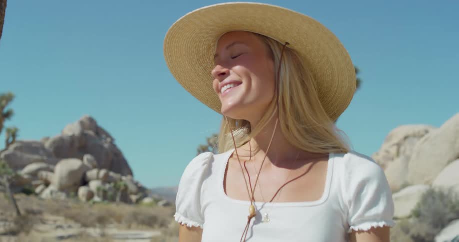 The video shows a woman with long blonde hair walking through a desert landscape she is wearing a straw hat a white top and brown shorts and she appears happy and relaxed the scenery includes large rocks and joshua trees under a clear blue s