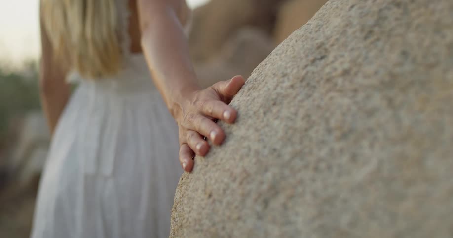 A womans hand rests on a large rock in a desert landscape with her back to the camera