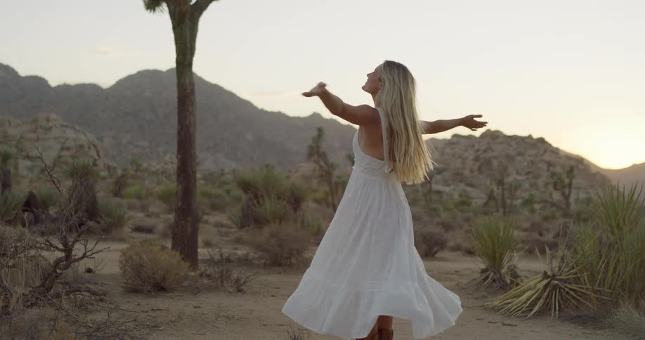 A woman with long blonde hair and a white dress stands on a dirt path in a desert with joshua trees and rocky hills turning around with her arms outstretched and smiling as she faces the camera