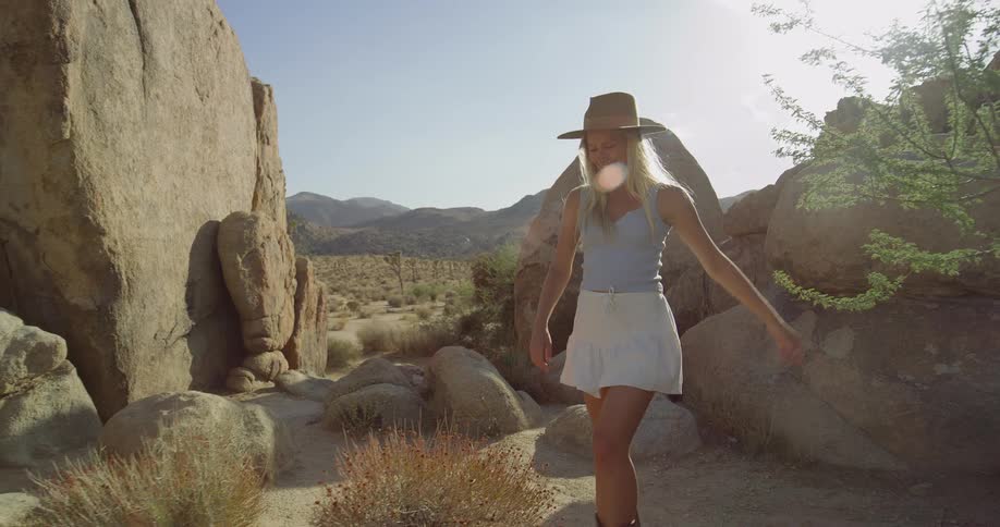 A woman with blonde hair wearing a beige hat a light blue tank top a white skirt and brown cowboy boots walks on a desert path with large rocks on both sides looking at the camera with a smile on her face