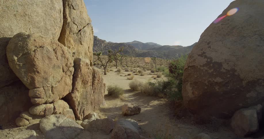 A camera pans up from a dirt path between large brown rocks to reveal a wide open desert landscape with sparse joshua trees and rolling mountains in the distance